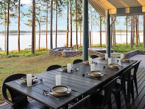 An outdoor dining area with a long table set for six, surrounded by chairs, overlooking a lake with trees and a stone fire pit in the background.