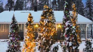 A log cabin with multiple Christmas trees adorned with lights, surrounded by snow.