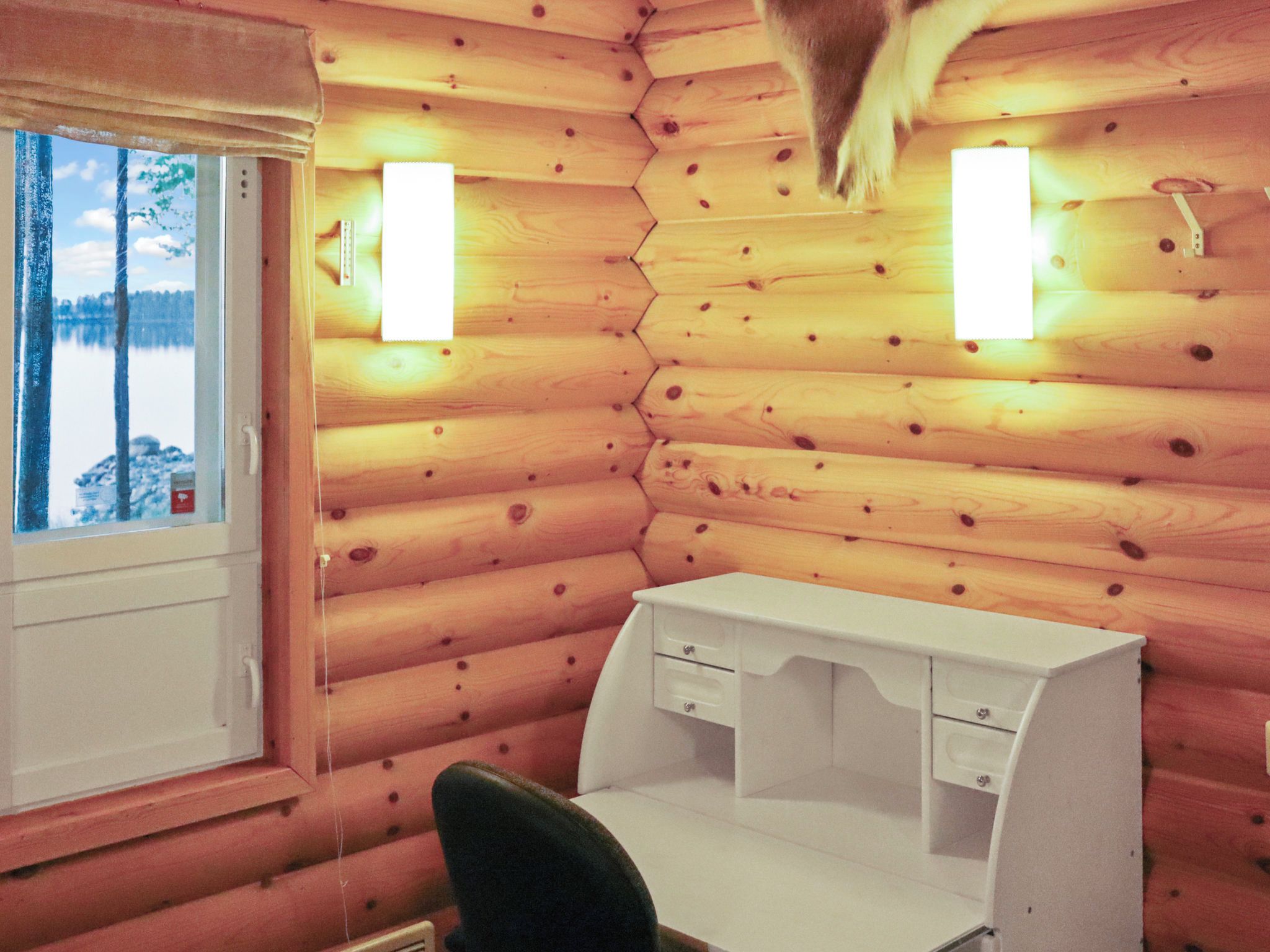 A cabin interior with a wooden log wall, a desk with drawers, a chair, and two wall-mounted lights.