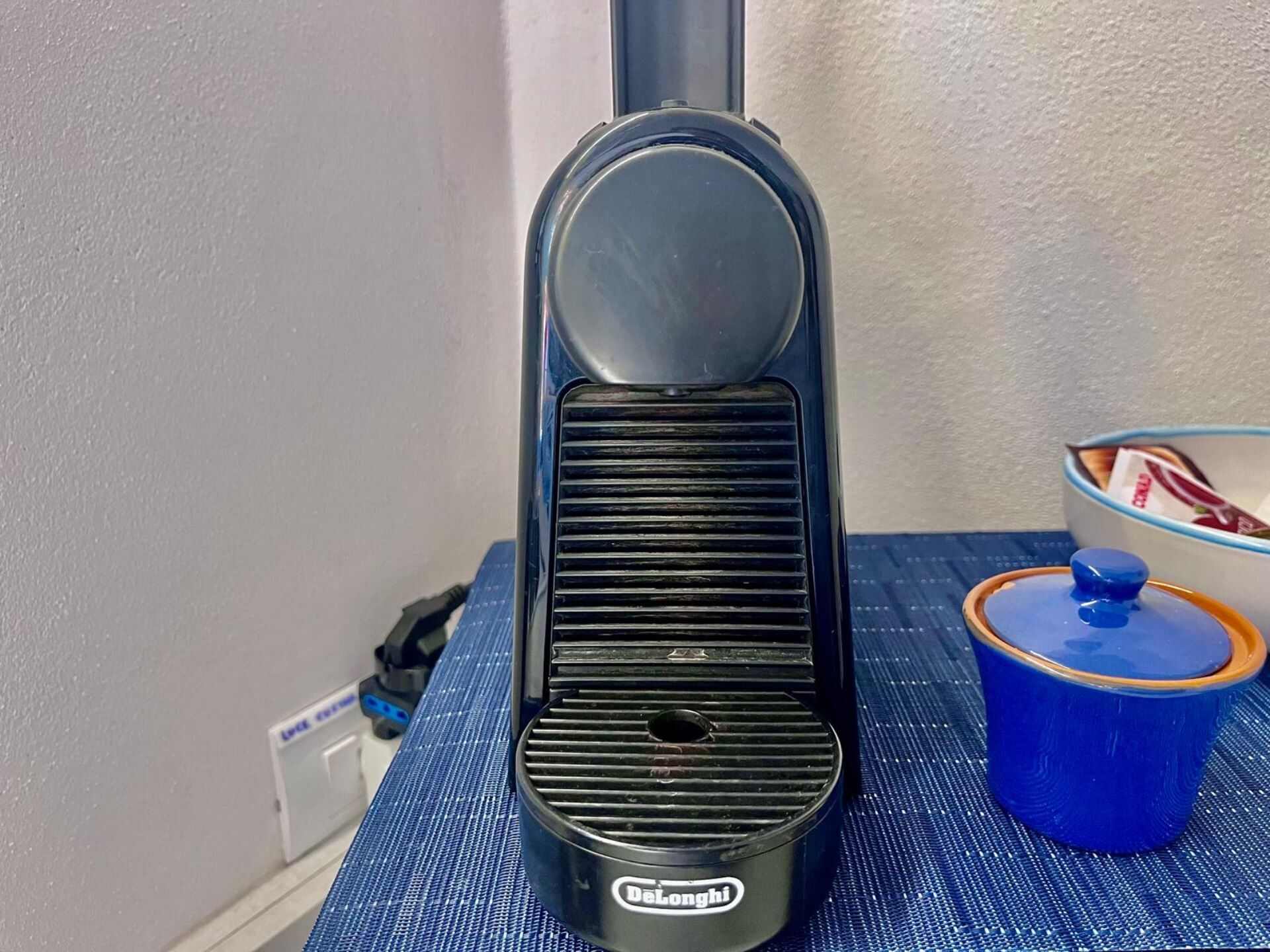 Une bouilloire électrique DeLonghi posée sur un tapis bleu, à côté d’une tasse bleue dotée d’un couvercle.