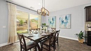 Un espace repas comprenant une grande table, des chaises et un lustre. Les murs sont ornés de peintures abstraites, et de grandes fenêtres offrent une vue sur l’extérieur.