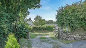 Un sentier rural mène à un mur de pierres et à une entrée fermée par un portail, le tout étant entouré de verdure, avec une maison visible au loin.