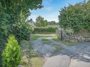 A rural pathway leading to a stone wall and a gated entrance, surrounded by greenery and a house in the background.