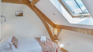 Une chambre avec un plafond en pente, un lit avec une literie blanche, une couvre-lit aux motifs colorés, une table de chevet avec une lampe et un tableau accroché au mur.