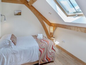 Une chambre avec un plafond en pente, un lit avec une literie blanche, une couvre-lit aux motifs colorés, une table de chevet avec une lampe et un tableau accroché au mur.