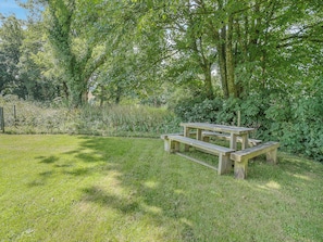A wooden picnic table in a grassy area with trees and bushes in the background.