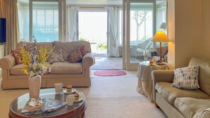 Ett vardagsrum med en beige soffa, ett soffbord i glas med tidningar, en vas med blommor och en lampa på ett sidobord.
