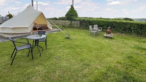Uma área de campismo com tendas, uma mesa e cadeiras, rodeada por um relvado e uma cerca de madeira.
