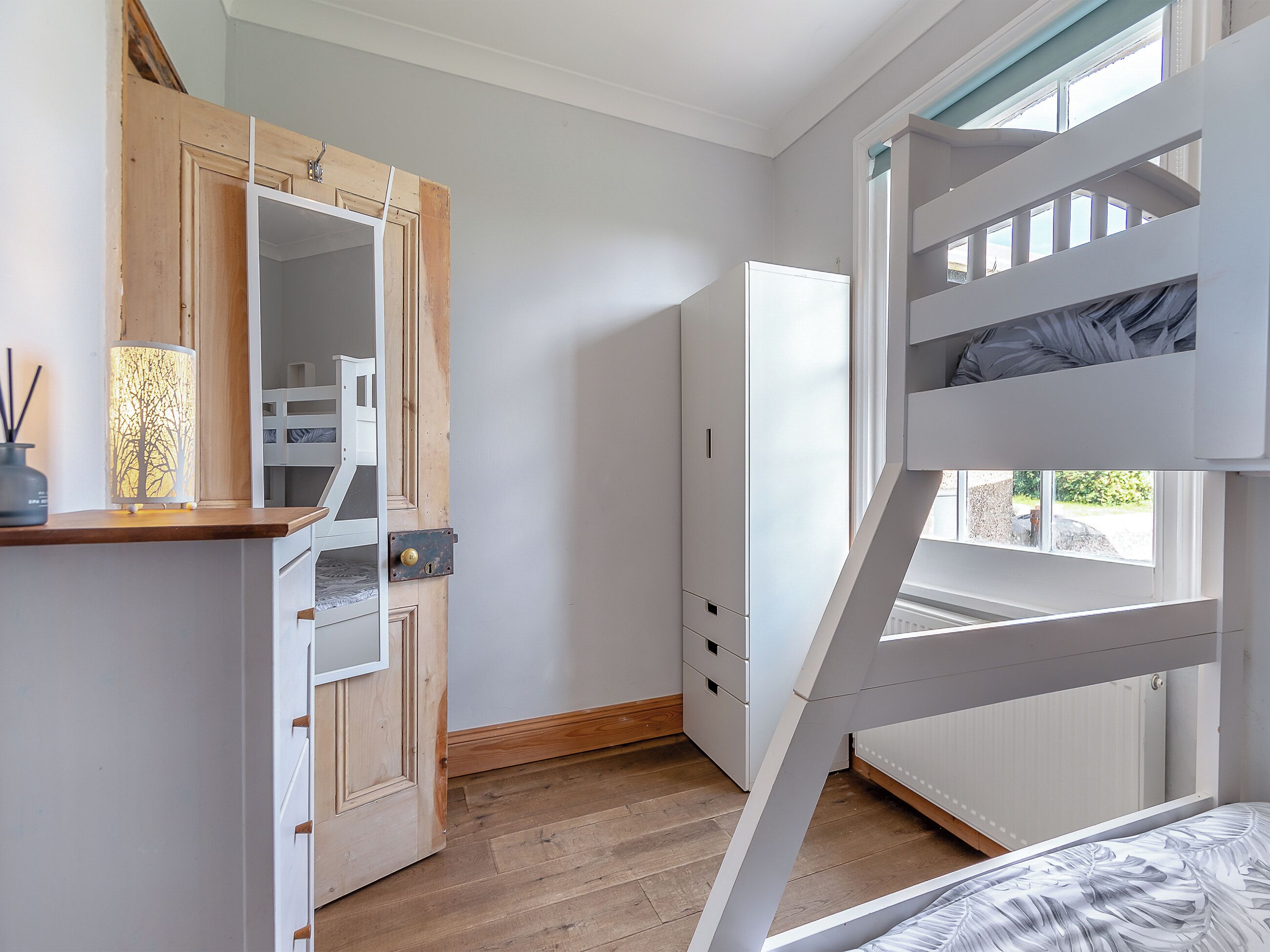 A bedroom with a bunk bed, a wooden wardrobe, a mirror, and a window.
