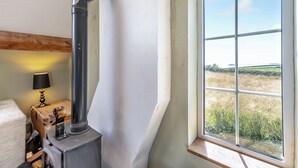 Una habitación con un ventanal grande, techo con vigas de madera, estufa negra y una pequeña mesa con lámpara.
