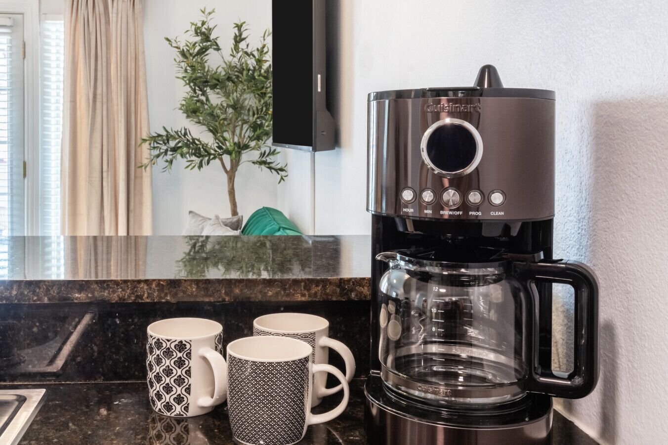 A coffee maker with a digital display, a glass carafe, and three patterned mugs on a kitchen counter.