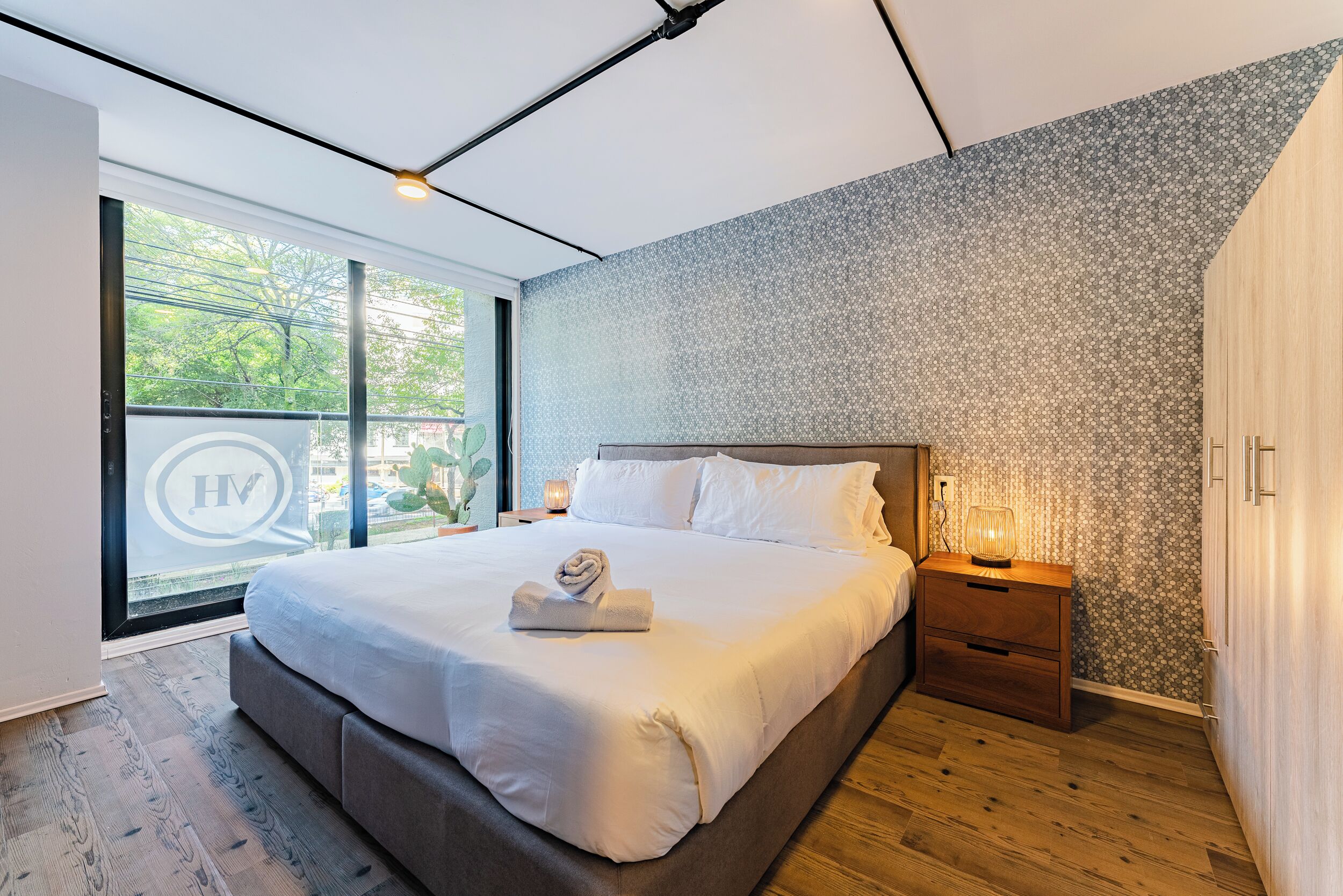 A modern bedroom with a large bed, a nightstand, a wooden wardrobe, and a view of greenery outside.