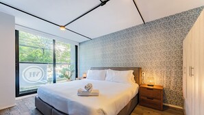 A modern bedroom with a large bed, a nightstand, a wooden wardrobe, and a view of greenery outside. - VH | Condesa 2 bedroom apartment | 245 (Ciudad de México)