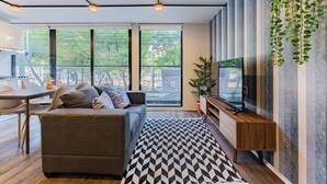 A modern living room with a sofa, a coffee table, and a TV on a wooden stand. There are large windows with a view of the outside. - VH | Condesa 2 bedroom apartment | 245 (Ciudad de México)