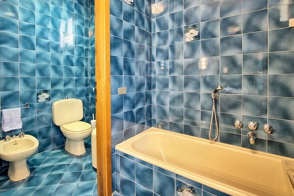Un baño con paredes de azulejos azules, un inodoro blanco, un bidé y una bañera con cabezal de ducha.