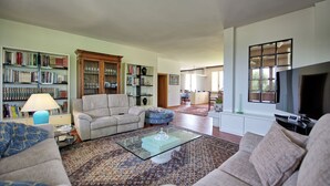 Una sala de estar con un sofá, un sillón, un reposapiés, una mesa de centro y un televisor. Se observan estanterías y un aparador con cristalería.