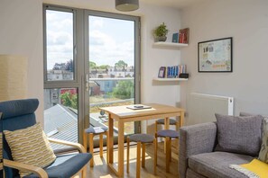 A modern living room with a large window, a wooden table with four chairs, a grey sofa, and a bookshelf with books and a framed picture.