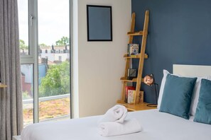 Una habitación de hotel con una cama, una estantería de madera con libros y vistas a edificios y árboles, además de una ventana con cortinas.