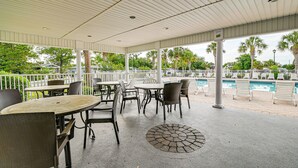 Un área de patio al aire libre con mesas y sillas, una piscina y palmeras al fondo.