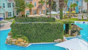 Una piscina con una isla rocosa, rodeada por un seto, y contigua a una hilera de casas coloridas.