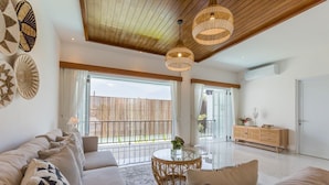 A spacious living room with a high ceiling, a wooden ceiling, a large sofa set, a round table with a vase, and a wooden cabinet.