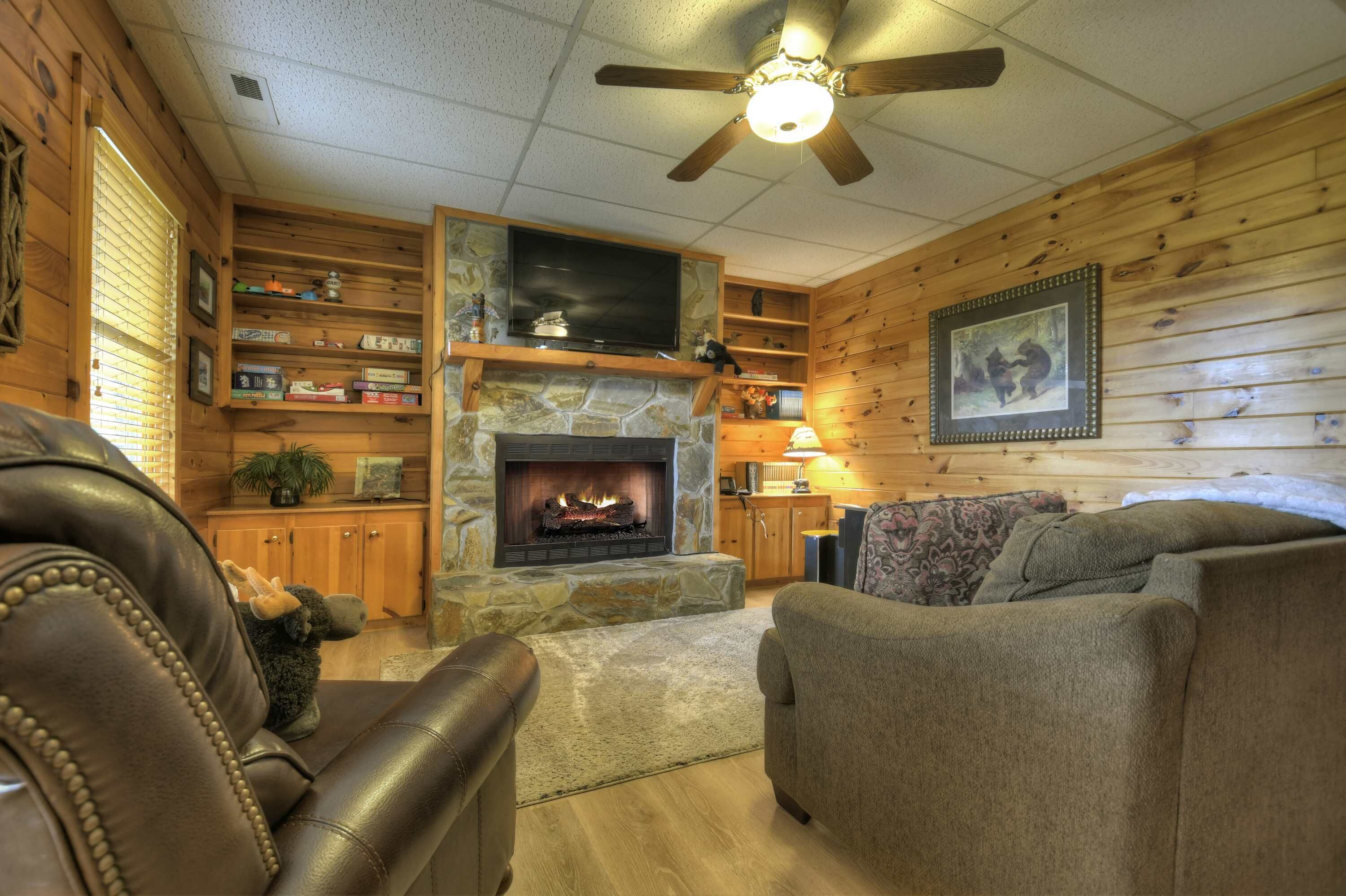 A cozy living room with a stone fireplace, leather furniture, wooden walls, and a ceiling fan.