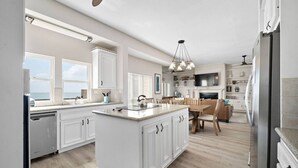 A modern kitchen with white cabinetry, a central island, and a dining area with a wooden table and chairs. - Bayview Home with Boat Slip & Stunning Views (Galveston)