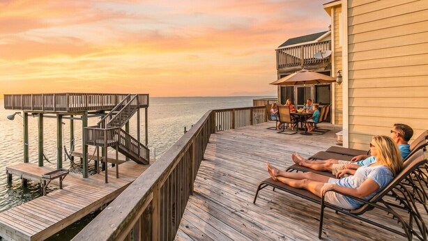 A wooden deck with a view of the water, a covered seating area, and a building with a balcony. - Bayview Home with Boat Slip & Stunning Views (Galveston)