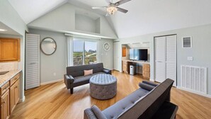 A spacious living area with a high ceiling, a ceiling fan, a large window with a view, a sofa, a round ottoman, and a kitchen area with cabinets and a TV. - Fisherman`s Dream: Game Room & Walk to Beach (Surfside)