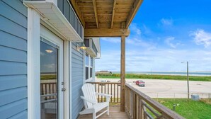 A wooden deck with a white chair, a table, and a view of a road and beach. - Fisherman`s Dream: Game Room & Walk to Beach (Surfside)