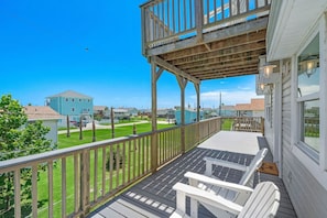 Una terraza de madera con baranda, una silla Adirondack blanca y vista a una zona residencial con casas y un cielo azul despejado.