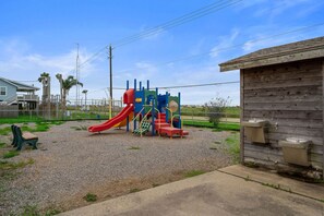 Ein Spielplatz mit einer roten Rutsche und Klettergerüsten, ein Holzgebäude mit Waschbecken sowie ein Haus im Hintergrund.