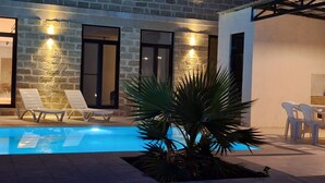 Una piscina con sillones de descanso y una palmera, rodeada de muros de piedra e iluminación moderna.