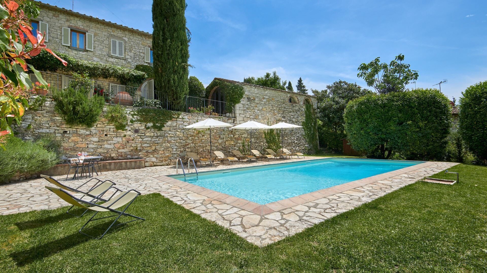 A swimming pool with lounge chairs and umbrellas, surrounded by stone walls and greenery.