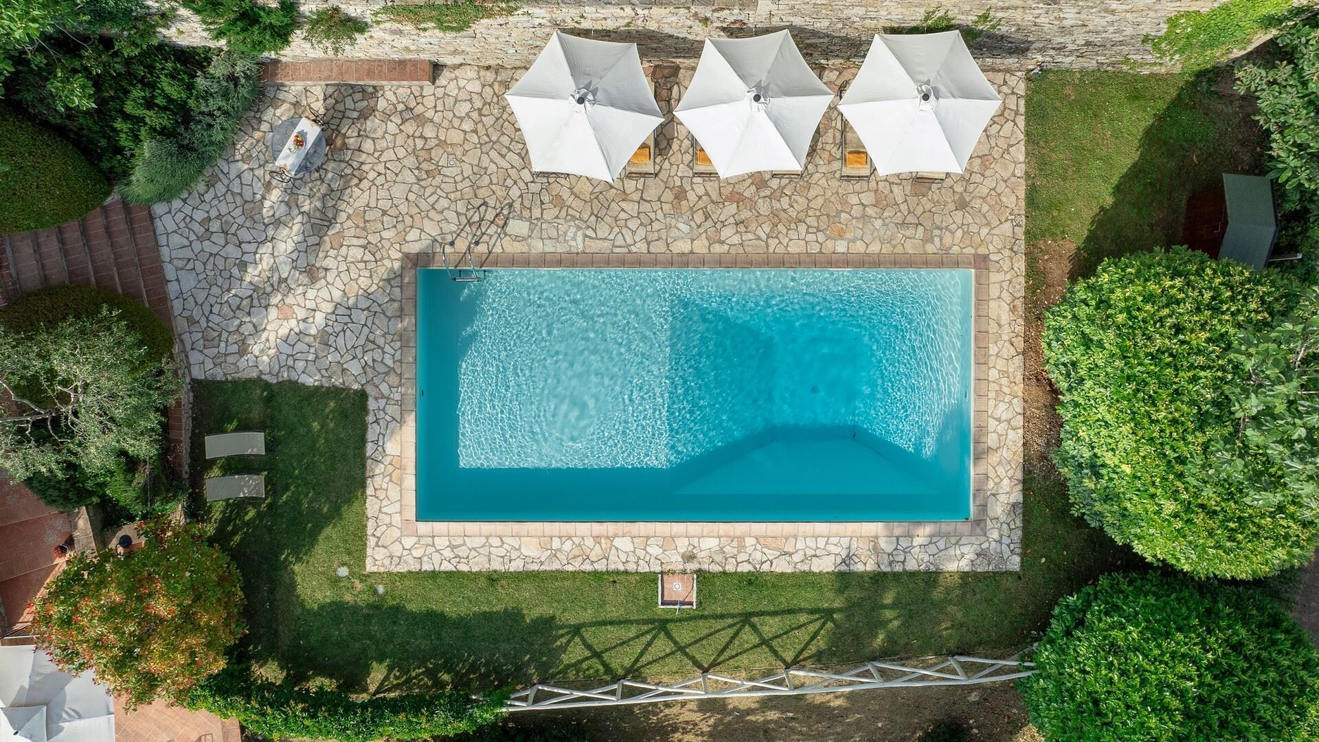 An aerial view of a rectangular swimming pool surrounded by stone tiles, with four white umbrellas and a stone wall.
