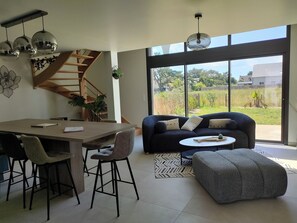 A modern living room with a dining area, a staircase, and large windows offering a view of the outdoors. - Invitation to the seaside Portbail 400m beach (Port-Bail-sur-Mer)