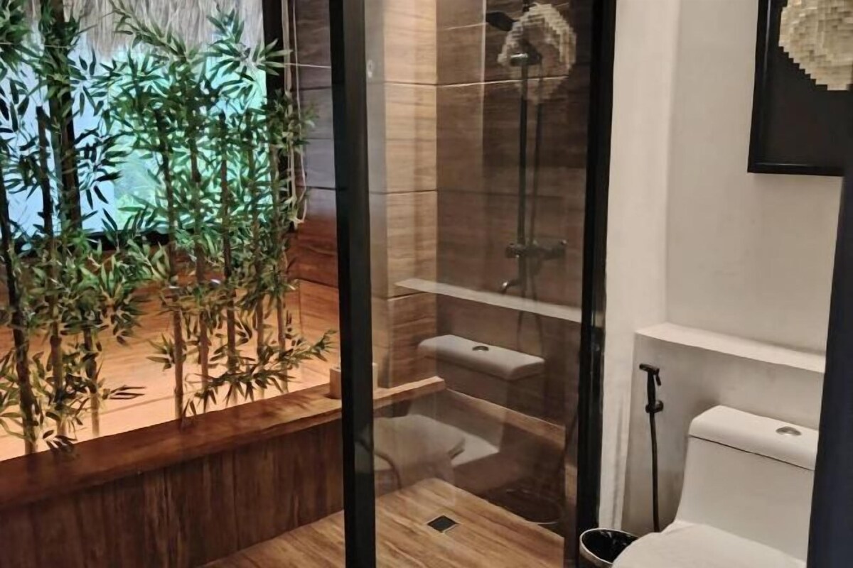 Baño con cabina de ducha de vidrio, inodoro y lavamanos. Hay una planta de bambú en el lado izquierdo.