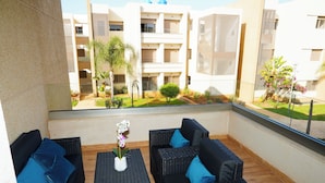 Un balcón con mobiliario de mimbre, una pequeña mesa con una flor y vistas a un edificio con varios balcones y vegetación.