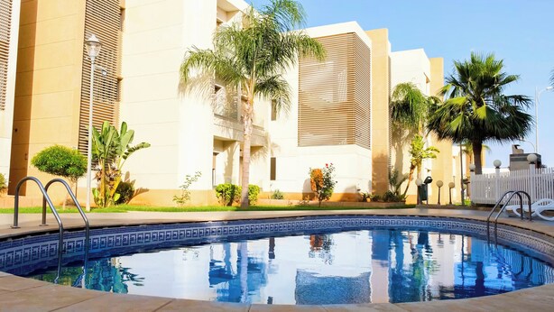 Una piscina con borde de azulejos, rodeada por una valla blanca, palmeras y un edificio de fachada color beige con listones verticales.