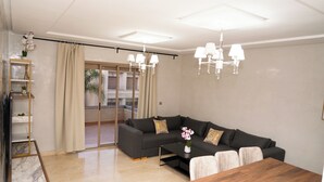 Una sala moderna con un sofá modular gris oscuro, una mesa de centro de madera y una mesa de comedor. Hay una puerta de vidrio que da a un balcón con vistas al exterior.