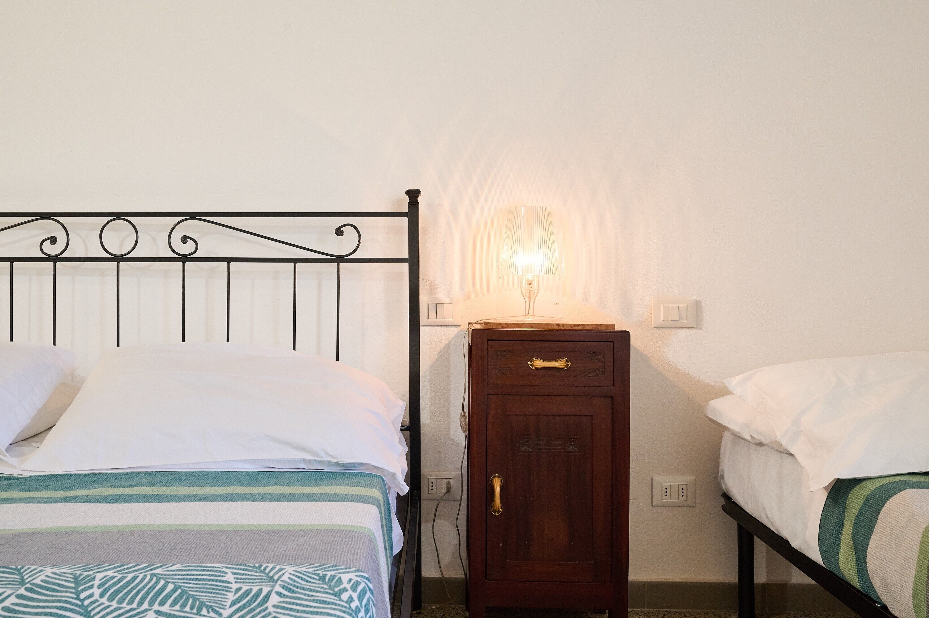Una cama individual con cabecera de metal, una mesita de noche con lámpara y un armario de madera.