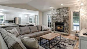 A modern living room with a stone fireplace, a gray sectional sofa, and a kitchen in the background.