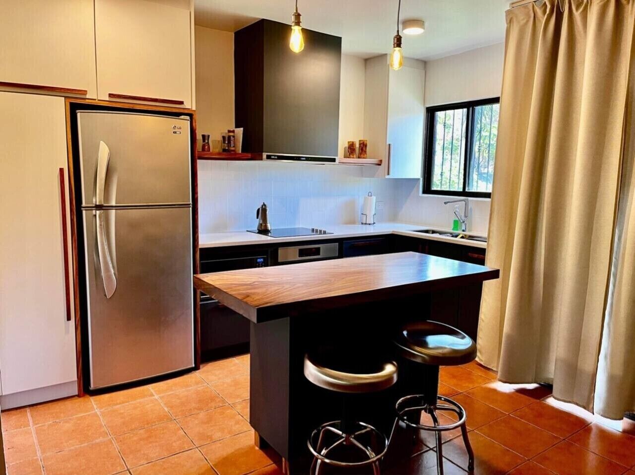 A modern kitchen with a stainless steel refrigerator, a wooden island with bar stools, and a window with curtains.