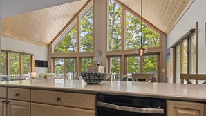 A kitchen with a large island, wooden cabinets, and a view of trees through large windows. - Attitude Adjustment on Glen Lake (Glen Arbor)