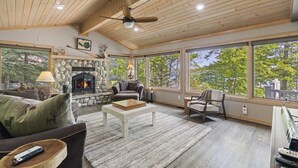 A spacious living room with a stone fireplace, large windows, and a ceiling fan. - Attitude Adjustment on Glen Lake (Glen Arbor)