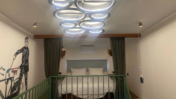 A hotel room with a modern ceiling light fixture, a bed with white bedding, and a green railing.