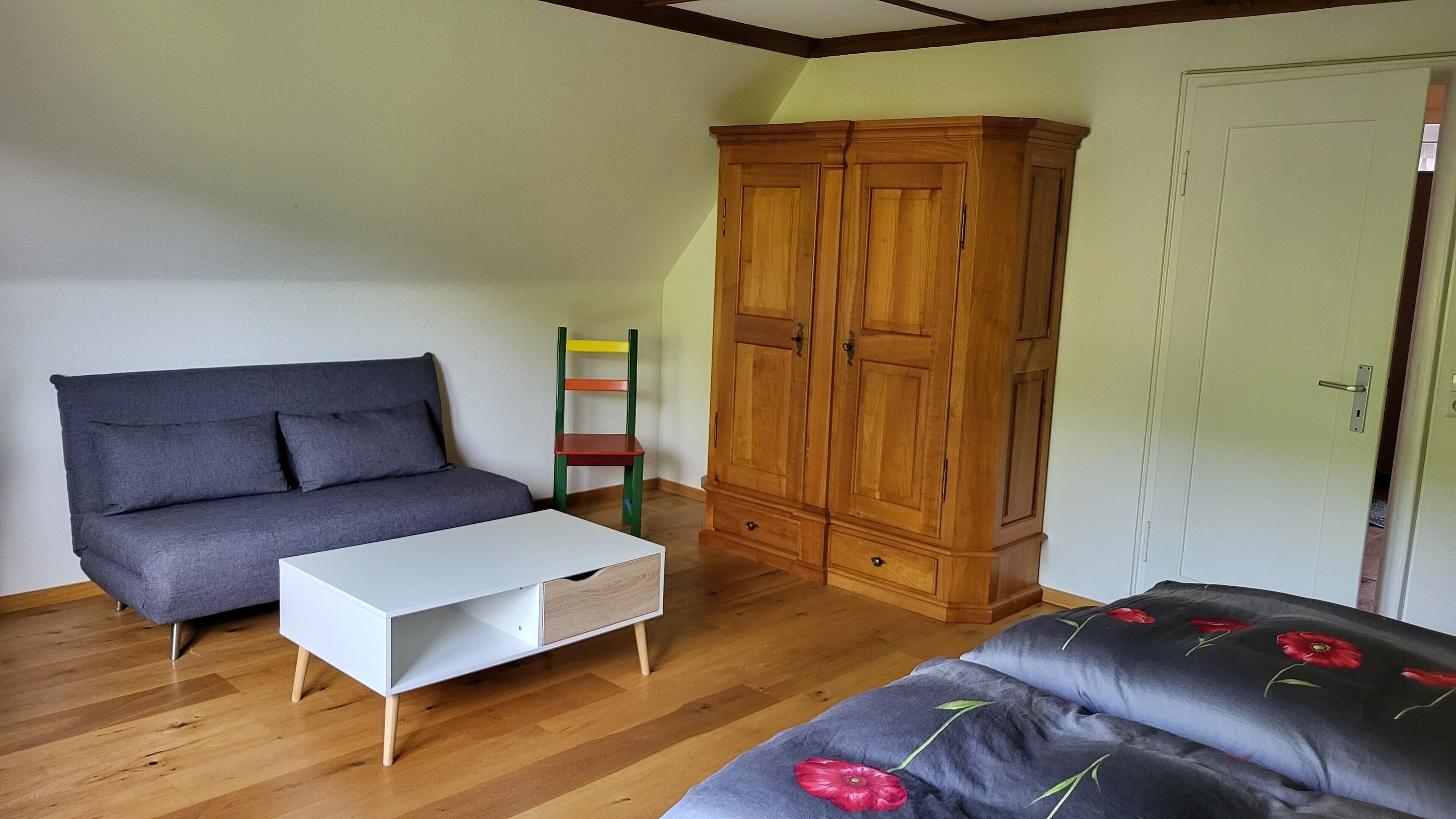Una stanza con un divano grigio, un tavolo bianco, un armadio in legno e un letto con copripiumino floreale.