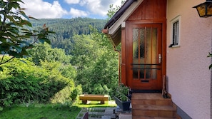 Une maison en bois avec une porte en verre, un banc et un jardin qui mène à une colline boisée.