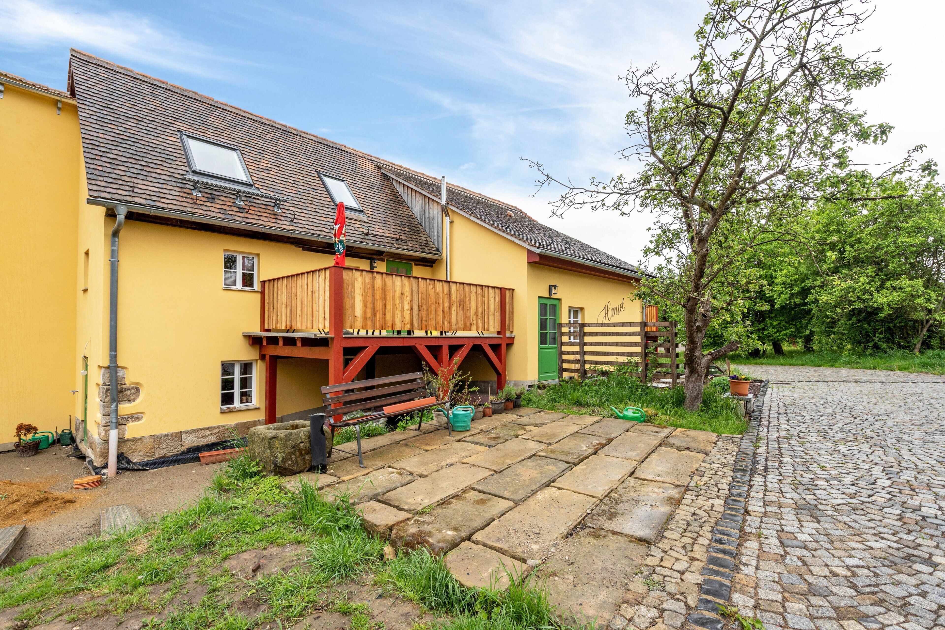 A yellow building with a wooden deck, a bench, and a tree.