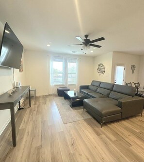 Un salon moderne comprenant un canapé d’angle, un téléviseur à écran plat, un ventilateur de plafond et un coin repas avec des chaises.